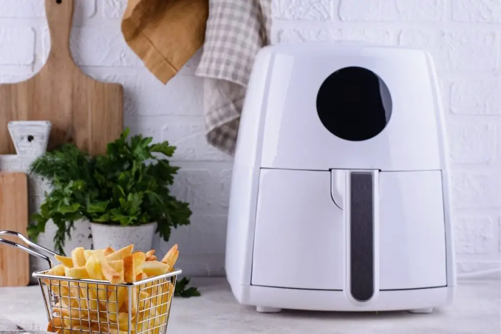 A white Air Fryer on a white counter with a basket of french fries next to it, and a cutting board behind it.