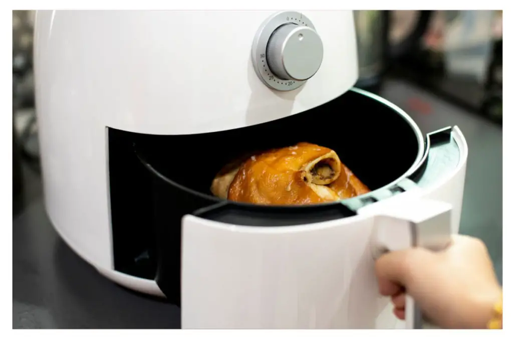 A white air fryer. a hand is visible pulling the door open and a roasted chicken is inside.