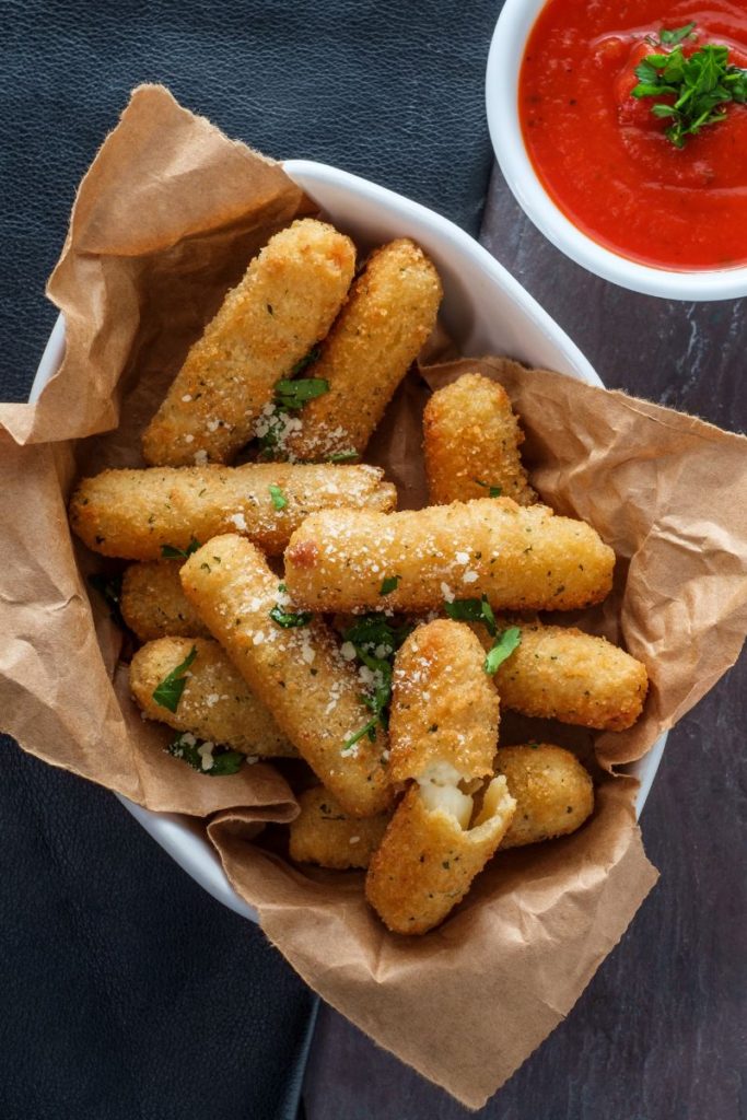Mozzarella Sticks in a white dish lined with brown paper. A small dish of marinara sauce next to it.