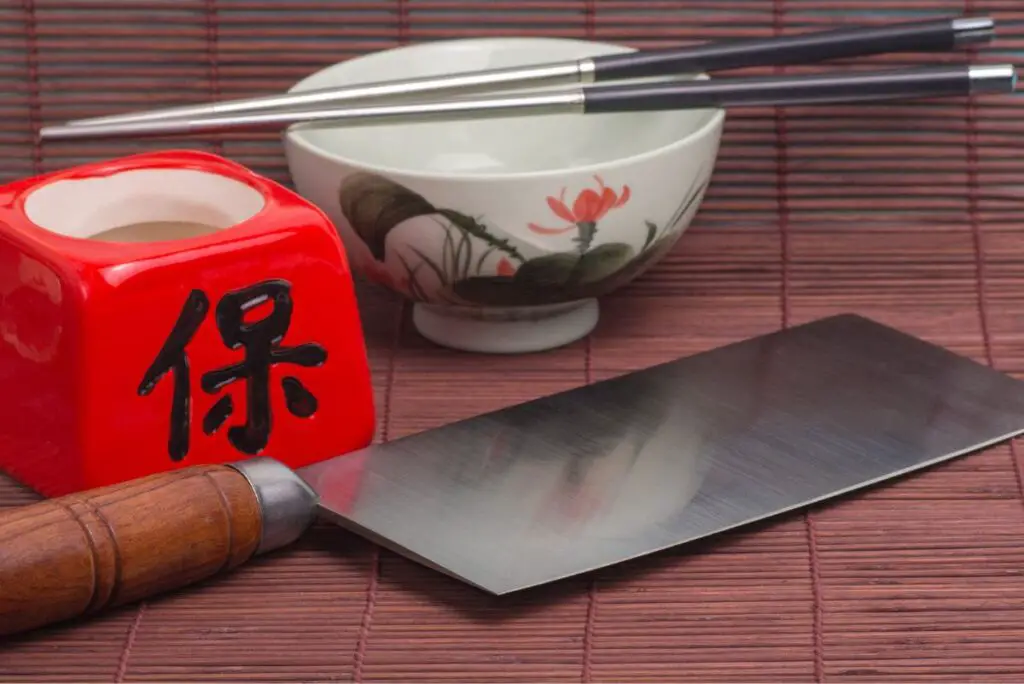 A Chinese cleaver on a wooden surface next to a red bowl with Chinese writing and a white bowl.