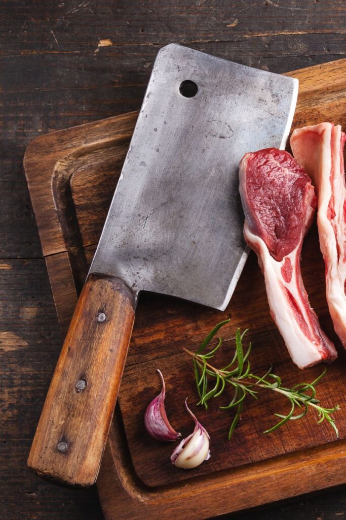A cleaver with a wooden handle, on a cutting board with raw lamb chops.