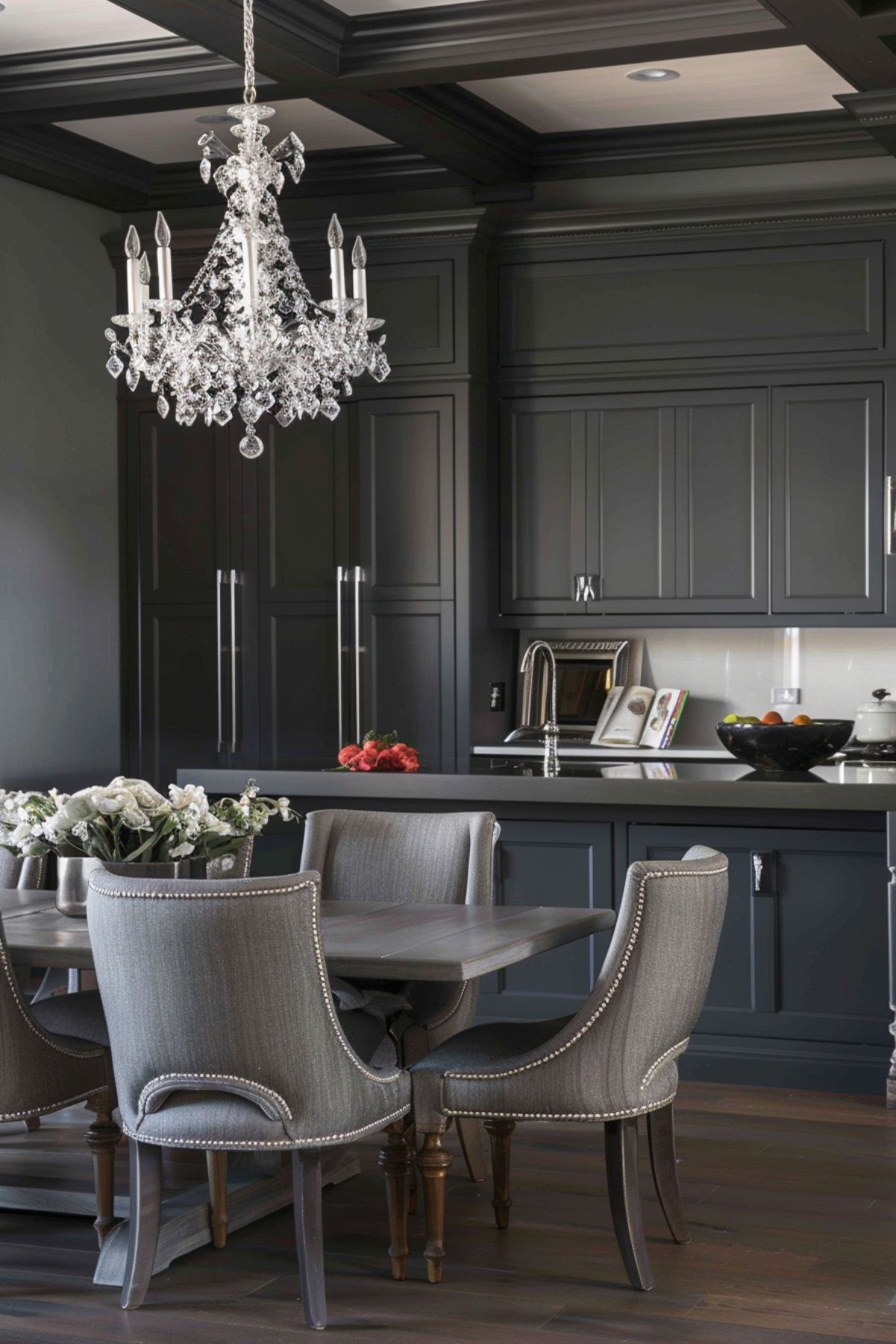 Sophisticated kitchen with dark cabinetry, a crystal chandelier, and a gray dining table with upholstered chairs. The kitchen features a sleek countertop, modern appliances, and elegant decor with fresh flowers, creating a luxurious and refined atmosphere.