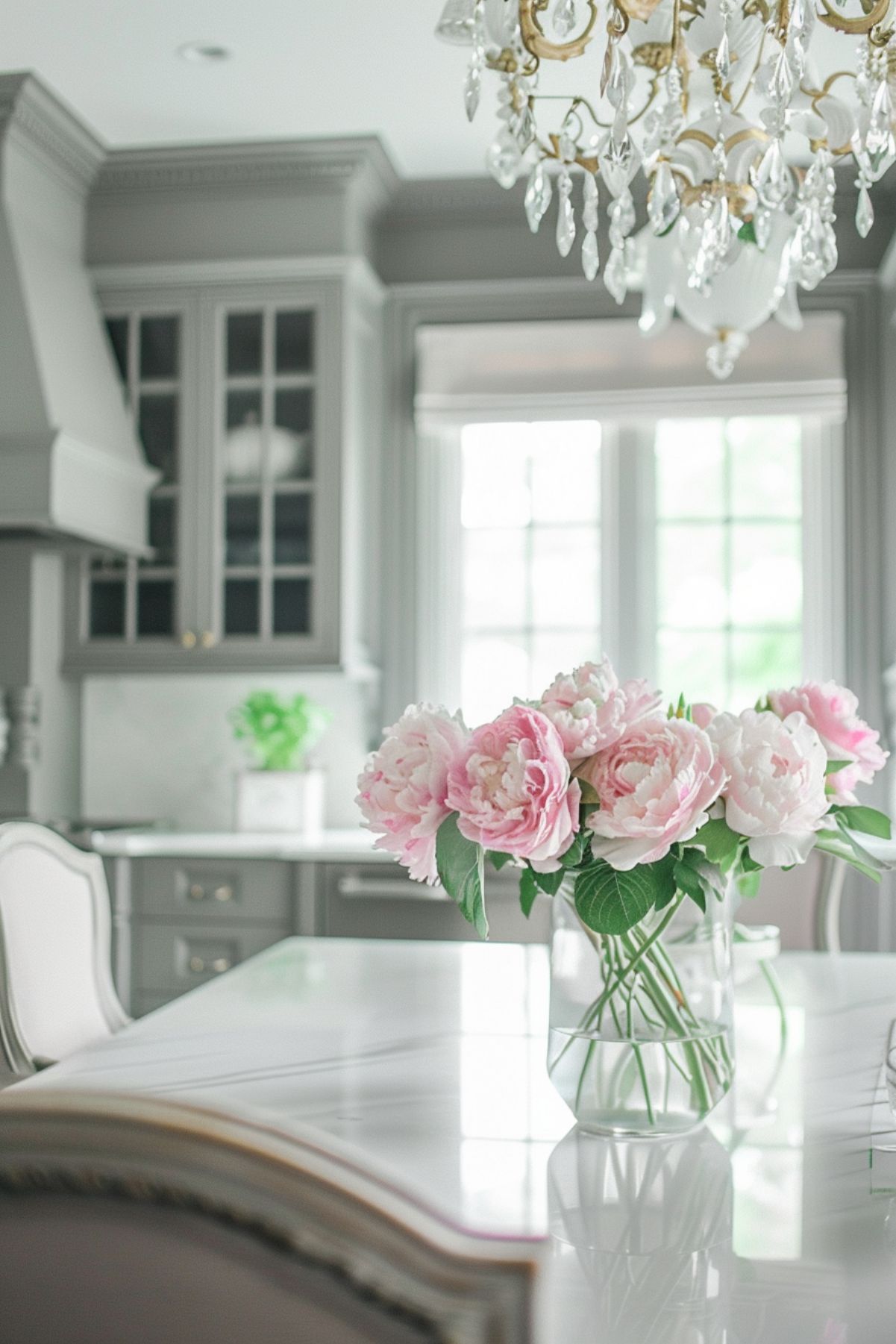 Elegant kitchen with light gray cabinetry and a marble countertop. The kitchen features a crystal chandelier, a vase of pink peonies, and large windows allowing natural light to flood the space, creating a bright and sophisticated atmosphere.