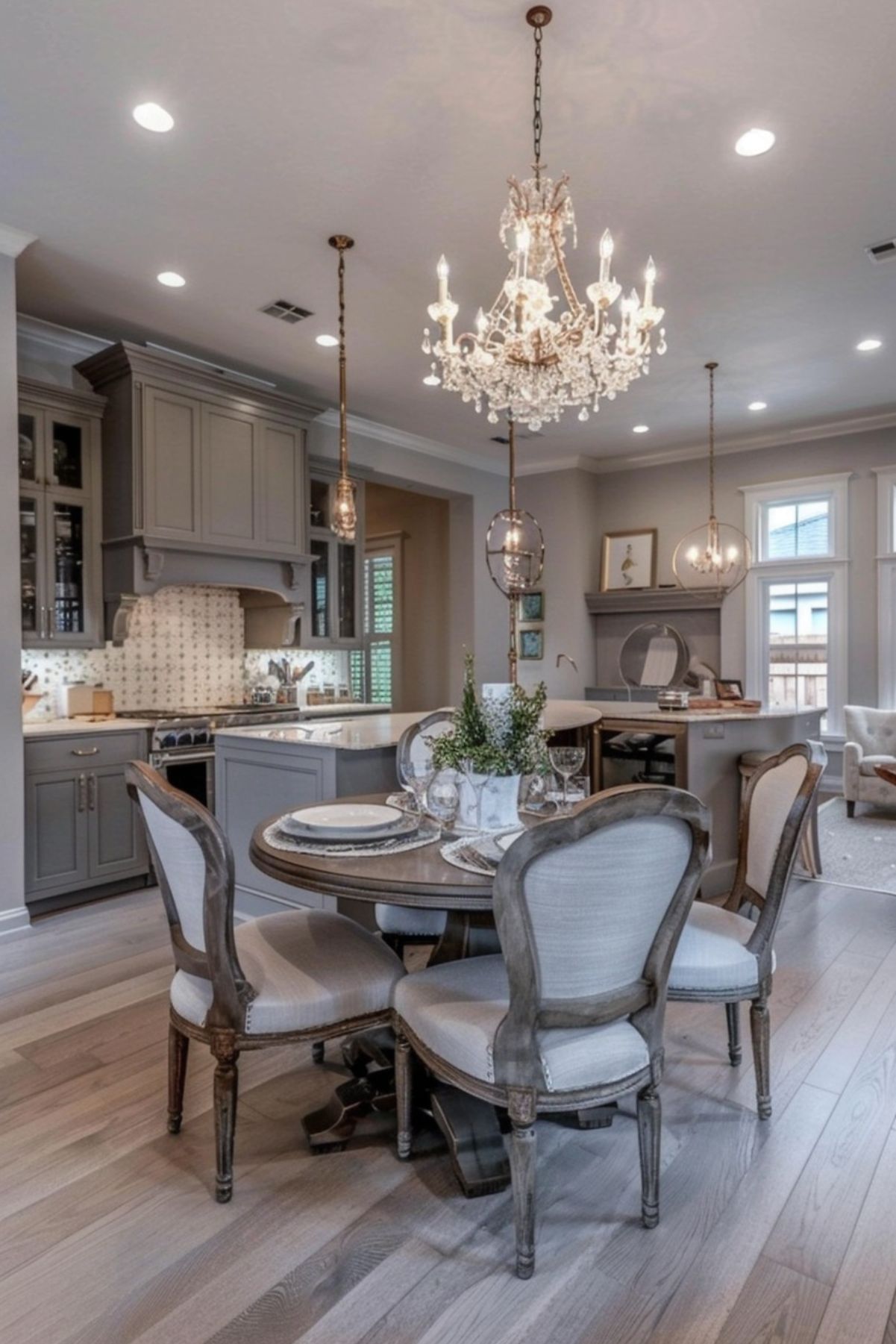 Elegant kitchen and dining area with gray cabinetry, a round wooden dining table, and upholstered chairs. The space features a crystal chandelier, pendant lights, and a mosaic tile backsplash, creating a refined and inviting atmosphere with modern and classic elements.