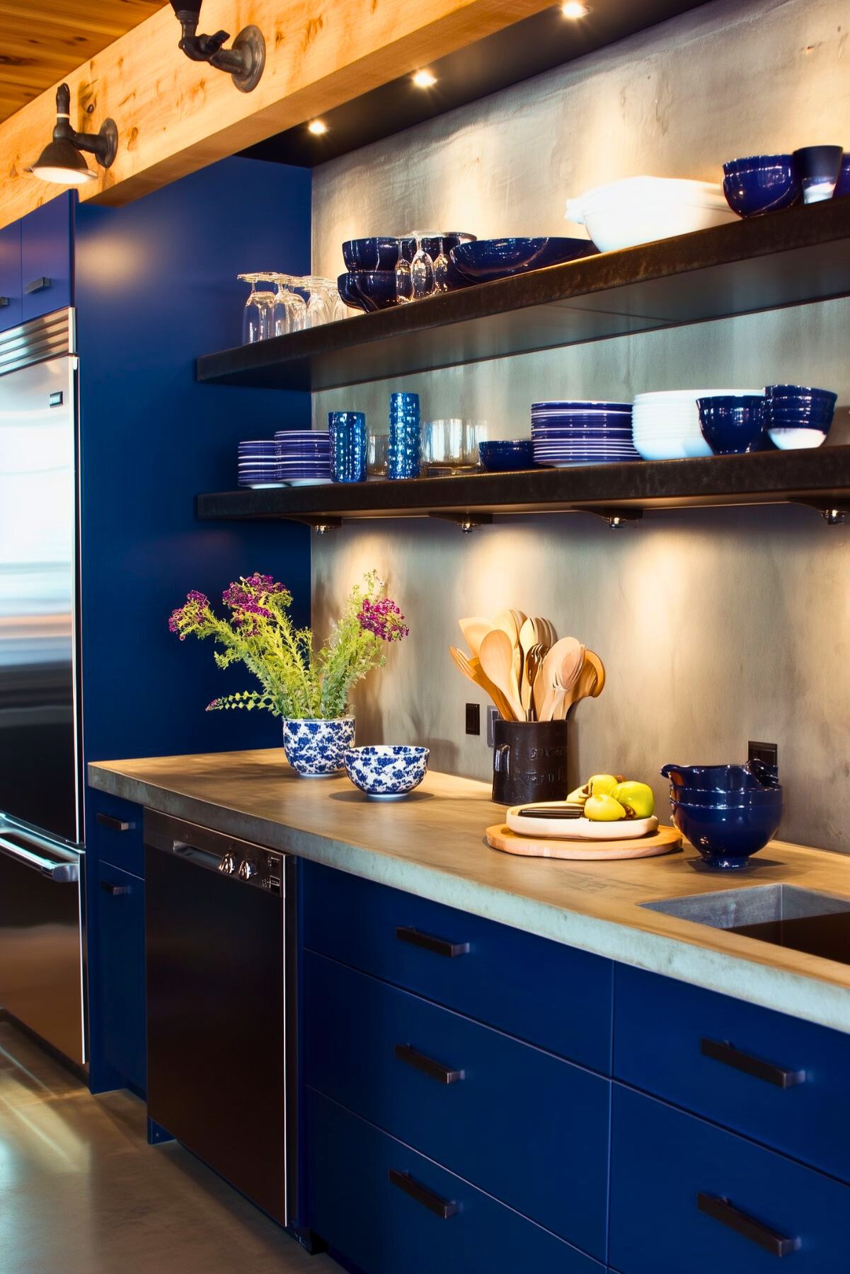 Contemporary navy blue kitchen with open shelving, wooden countertop, and under-shelf lighting; features vibrant blue and white dishware, decorative plants, and a modern industrial aesthetic.