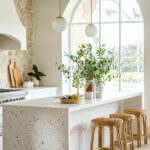 A bright, modern kitchen with a terrazzo-style white island featuring speckled patterns and wooden bar stools with woven seats. The space is decorated with greenery in minimalist vases, large arched windows bringing in natural light, and a stone accent wall behind a sleek white stove.