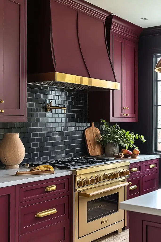 Stylish kitchen with deep burgundy cabinetry and brass hardware, featuring a bold range hood with a brass trim. The backsplash is covered in glossy black subway tiles, complementing the gold-tone stove and matching accents. Decorative elements include a wooden cutting board, a vase with greenery, and fresh fruit on the countertop, adding natural touches to the modern design.