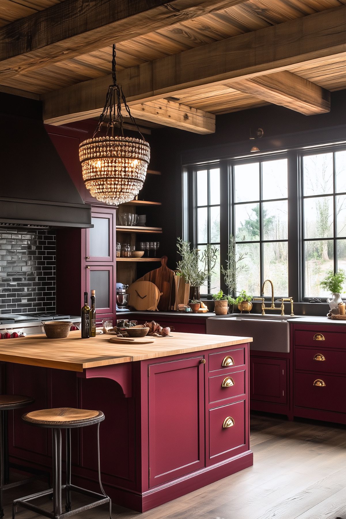 Rustic kitchen with burgundy cabinetry and brass hardware, featuring a large wooden island with industrial-style bar stools. A farmhouse sink sits beneath a trio of large windows, allowing natural light to flood the space. The kitchen includes a black subway tile backsplash, a decorative crystal chandelier, and exposed wooden beams on the ceiling. Open shelving holds kitchen essentials, while potted herbs and olive oil bottles add to the natural, cozy atmosphere.