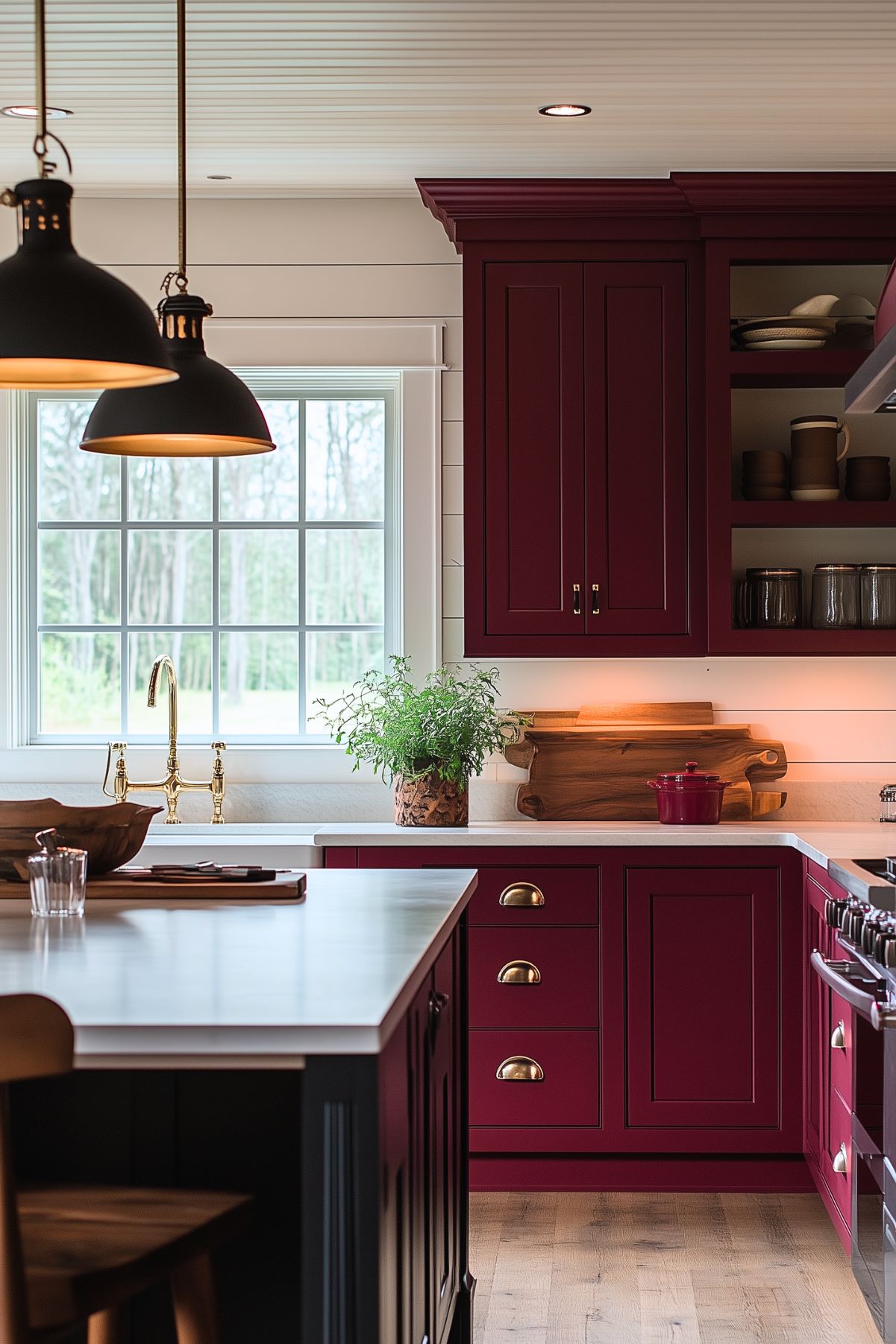 Farmhouse-style kitchen with burgundy cabinets and brass hardware, featuring a large window above the sink that provides natural light. The space includes white countertops, a wooden cutting board, and open shelving displaying ceramic dishes. Black pendant lights hang above the island, which is topped with a wooden serving bowl and cutting boards. A potted plant adds a touch of greenery to the cozy, rustic kitchen.