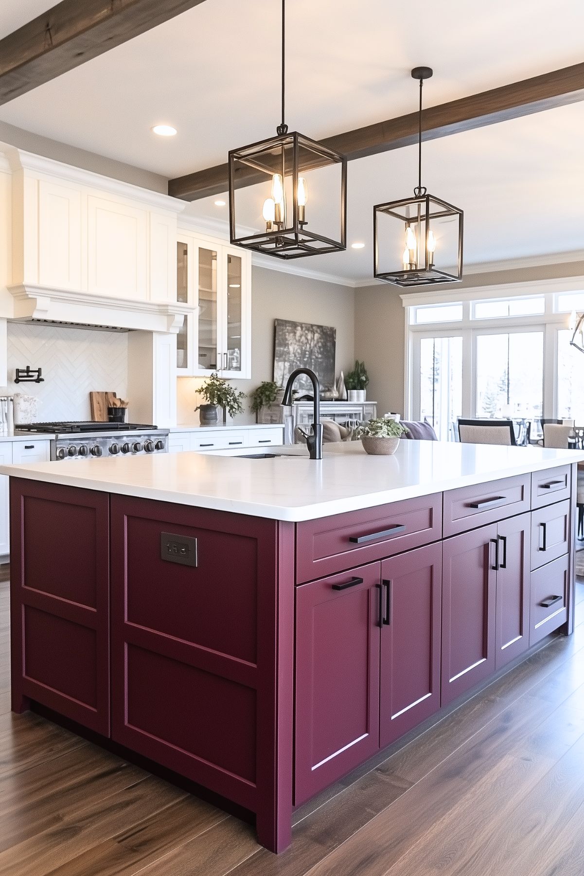 Modern kitchen featuring a large burgundy island with white countertops and black hardware, accented by two geometric pendant lights hanging above. The kitchen has white cabinetry and a stainless steel gas stove, with a herringbone backsplash adding texture. Dark wood beams on the ceiling complement the hardwood flooring, while natural light floods the space through large windows in the adjacent dining area.