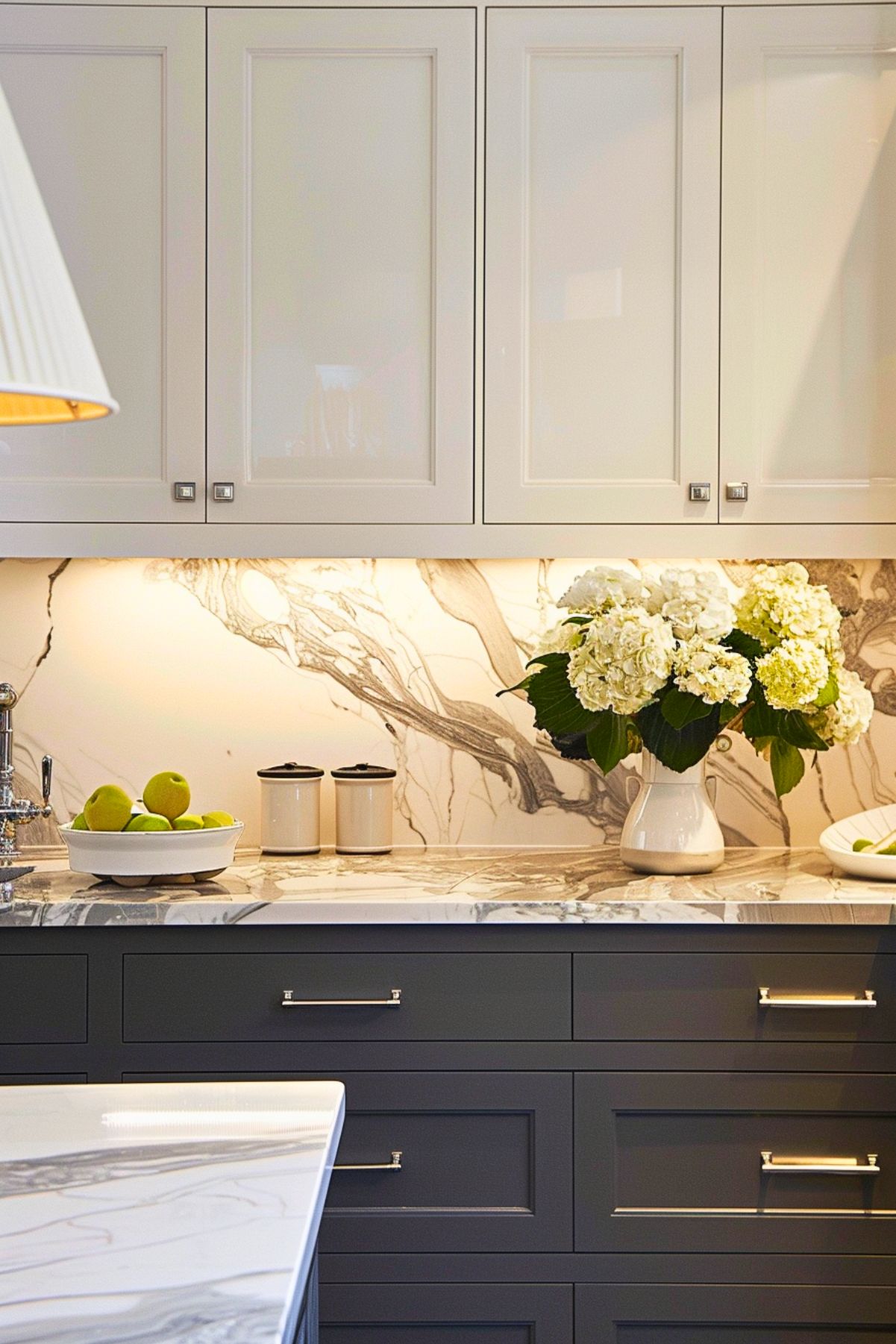 Stylish kitchen with glossy cream upper cabinets and charcoal grey lower cabinets, featuring a striking marble backsplash with bold veining that complements the marble countertop. The counter is decorated with a bowl of green apples, ceramic canisters, and a vase filled with white hydrangeas, adding a fresh and elegant touch to the sleek, modern design.