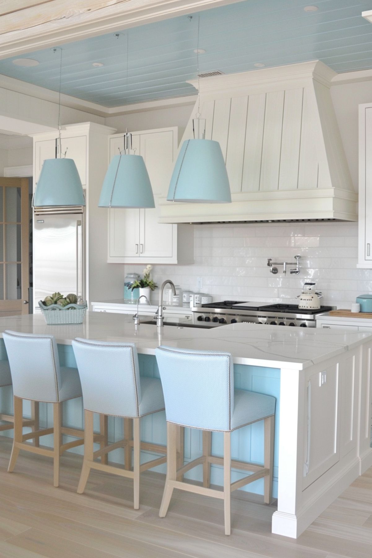 A coastal-inspired kitchen featuring white cabinetry and light blue accents. The space includes a large island with a white countertop and light blue barstools. Above the island, three blue pendant lights hang from a white shiplap ceiling with light blue insets. The kitchen also showcases stainless steel appliances and a decorative range hood, contributing to the airy and serene beach-house vibe.