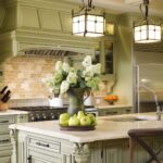 A beautifully designed kitchen showcasing a blend of olive green cabinetry and cream accents. The centerpiece is a large island with intricate detailing and a smooth, light-colored countertop, adorned with a vase of fresh flowers and a tray of green apples. The backsplash features warm-toned brick tiles, adding a rustic charm to the space. Modern stainless steel appliances complement the traditional cabinetry, and elegant pendant lights with wrought iron details hang above the island, providing a warm and inviting glow. The overall design combines classic elements with modern functionality, creating a welcoming and stylish kitchen.