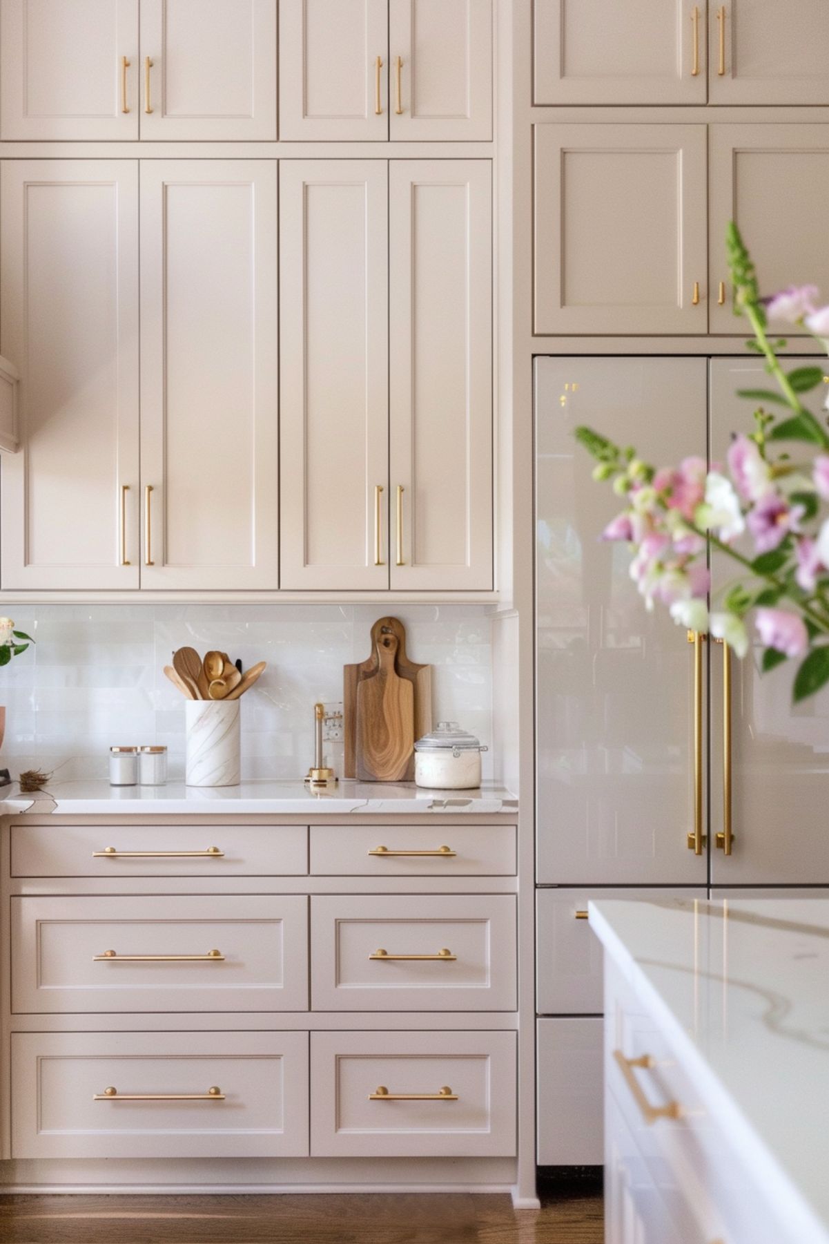 A stylish kitchen with blush beige cabinets and gold handles. The tall cabinets offer ample storage. A marble countertop holds a utensil holder, cutting board, and canister. The backsplash features subtle white tiles. A built-in refrigerator with matching cabinetry and gold handles blends seamlessly into the design. A flower arrangement adds a touch of natural beauty.