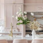 A stylish kitchen with blush beige cabinets and a white marble countertop island. The island is paired with plush pink bar stools with gold bases. A gold faucet stands out on the island, which is decorated with vases of fresh flowers. The backsplash and range hood feature marble and gold accents, enhancing the elegant design.