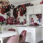 Contemporary kitchen with dark floral wallpaper featuring large red and pink flowers. The space includes white cabinets, a white kitchen island with a sink, gold fixtures, pink velvet chairs, a white round dining table, and modern pendant lights.