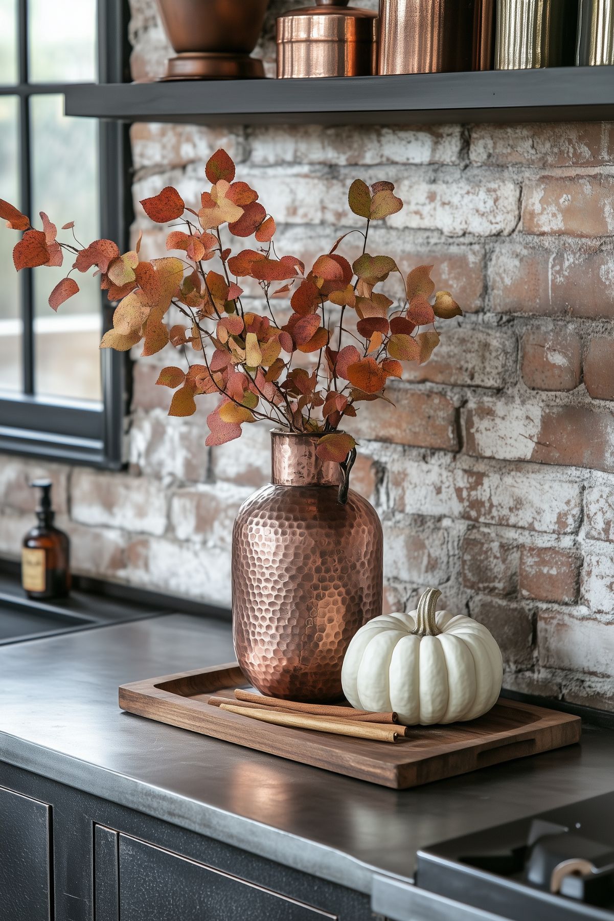 A modern industrial kitchen with fall decor featuring a hammered copper vase filled with autumn leaves. A white pumpkin and cinnamon sticks rest on a wooden tray, placed on a sleek stainless steel countertop. The exposed brick wall and dark cabinetry enhance the rustic yet contemporary feel of the space, creating a stylish autumn ambiance.