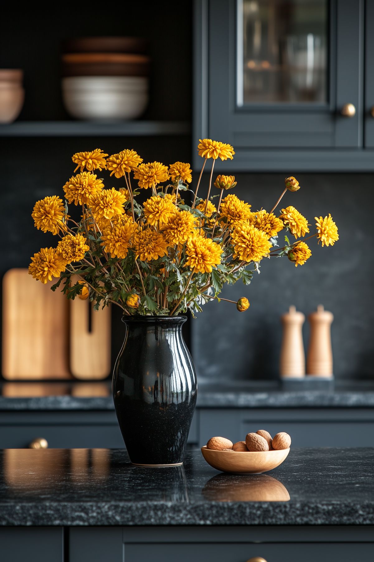 A stylish kitchen counter featuring a black vase filled with bright yellow chrysanthemums, creating a striking contrast against the dark cabinetry and countertop. A small wooden bowl with walnuts adds a natural touch, enhancing the sophisticated and warm autumn ambiance of the space.