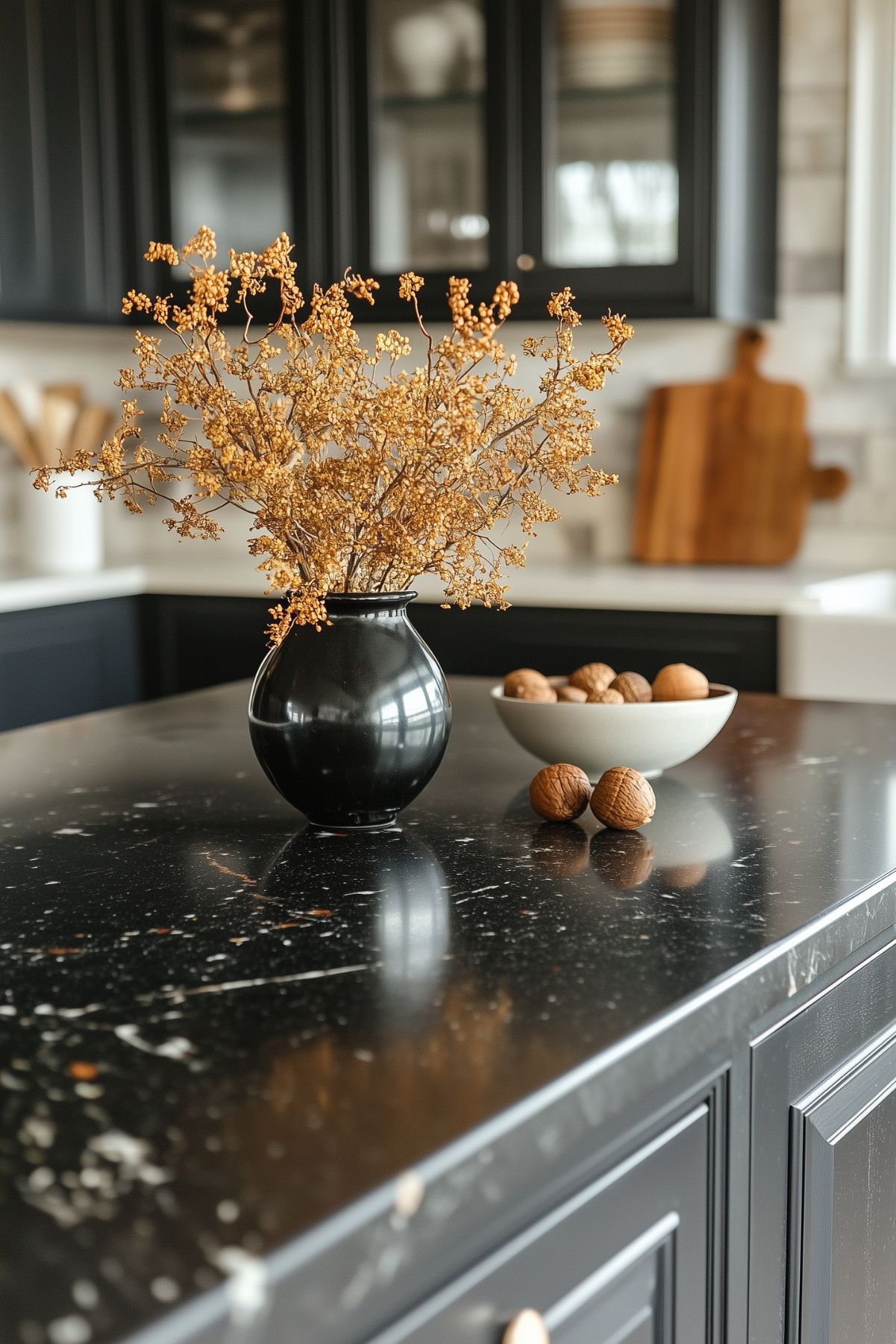 A sleek kitchen with dark cabinetry and a black marble countertop, featuring a black vase with dried autumn branches. A white bowl filled with walnuts adds a natural touch to the elegant and minimalist fall decor. The background includes cutting boards and kitchen utensils, complementing the modern aesthetic.