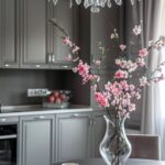 Elegant gray kitchen with a crystal chandelier hanging above a round table. A glass vase filled with blooming pink cherry blossoms is the centerpiece on the table, adding a pop of color to the modern, monochromatic decor. White tableware is neatly arranged on the table, and the background features sleek gray cabinets with metallic handles, creating a sophisticated and calm atmosphere.