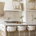 A sophisticated kitchen with a white island, brass accents, and ivory cabinetry. The space features brass lantern-style pendant lights, plush bar stools, and a herringbone tile backsplash.