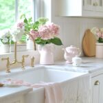 A bright kitchen with a farmhouse sink featuring a brass faucet. The windowsill is adorned with pink and white hydrangeas in vases, while lace curtains add a delicate touch. A pink teapot and other pastel kitchen decor items complete the vintage-inspired look.