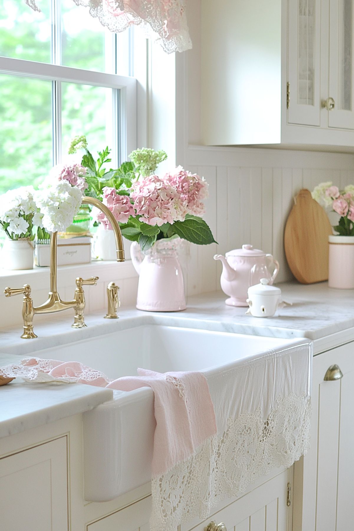 A bright kitchen with a farmhouse sink featuring a brass faucet. The windowsill is adorned with pink and white hydrangeas in vases, while lace curtains add a delicate touch. A pink teapot and other pastel kitchen decor items complete the vintage-inspired look.