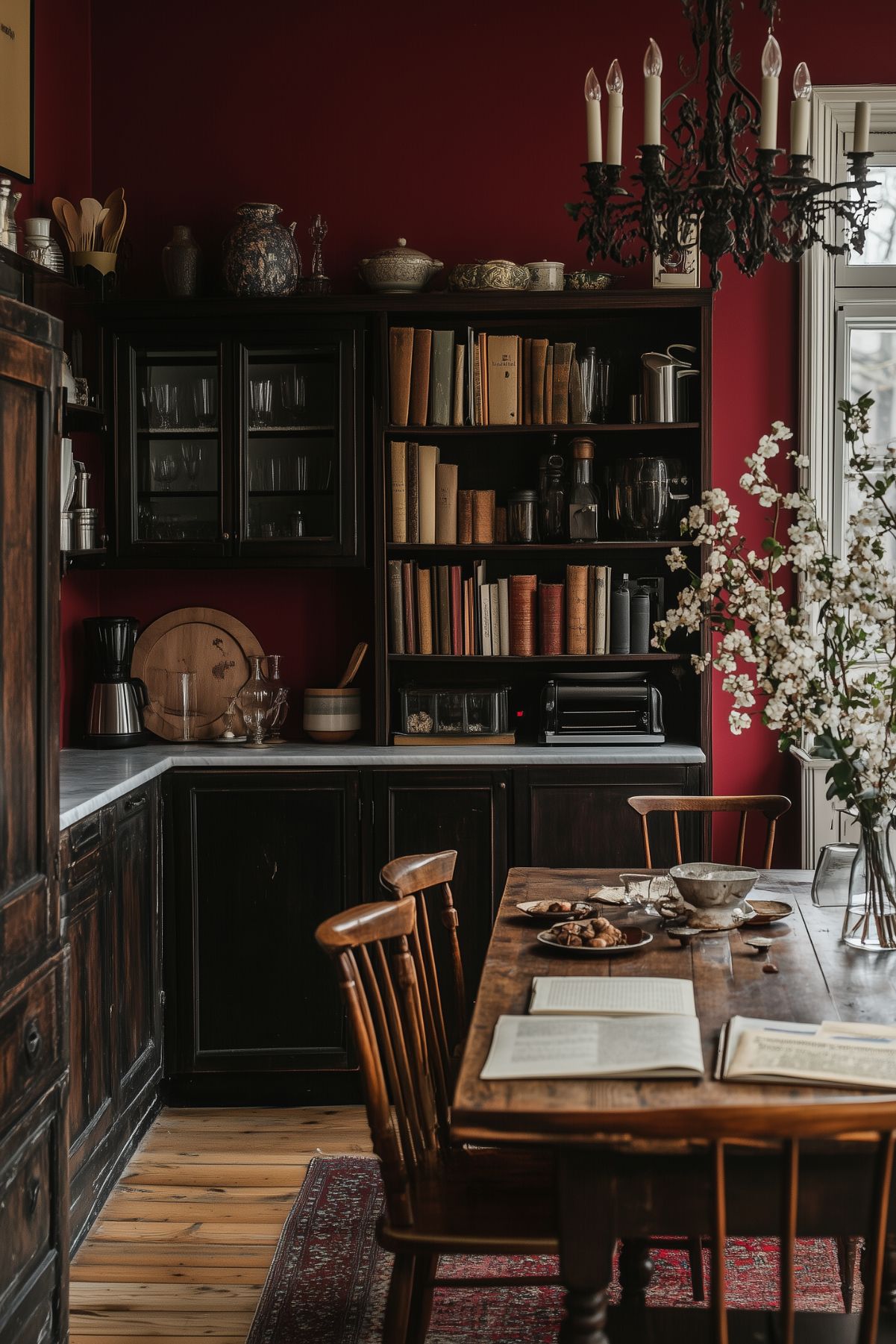 A warm, dark kitchen with deep red walls and dark wooden cabinetry. Shelves are filled with vintage books and kitchenware, while the countertop holds a variety of small appliances and utensils. A rustic wooden dining table is in the foreground, partially set with plates of pastries, open books, and a floral centerpiece in a vase. A black iron chandelier adds a touch of old-world charm, enhancing the dark academia aesthetic. The room is softly lit by natural light coming through a nearby window.