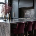 Modern kitchen with sleek dark cabinetry and a marble-textured island. The gold hardware and fixtures add a touch of luxury, while deep purple velvet bar stools bring a pop of color and warmth. A large vase with burgundy branches sits on the island, softening the minimalist design with a natural element.