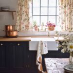 A quaint kitchen featuring dark lower cabinets with brass hardware, a farmhouse sink, and a butcher block countertop. The window is adorned with floral curtains, letting in natural light, and potted plants add a fresh touch to the windowsill. Copper pots rest on the counter, enhancing the rustic, country feel. An old wooden dining table with mismatched chairs sits in the foreground, creating a cozy and lived-in atmosphere. The exposed wooden beam above the window adds a charming rustic detail to the space.