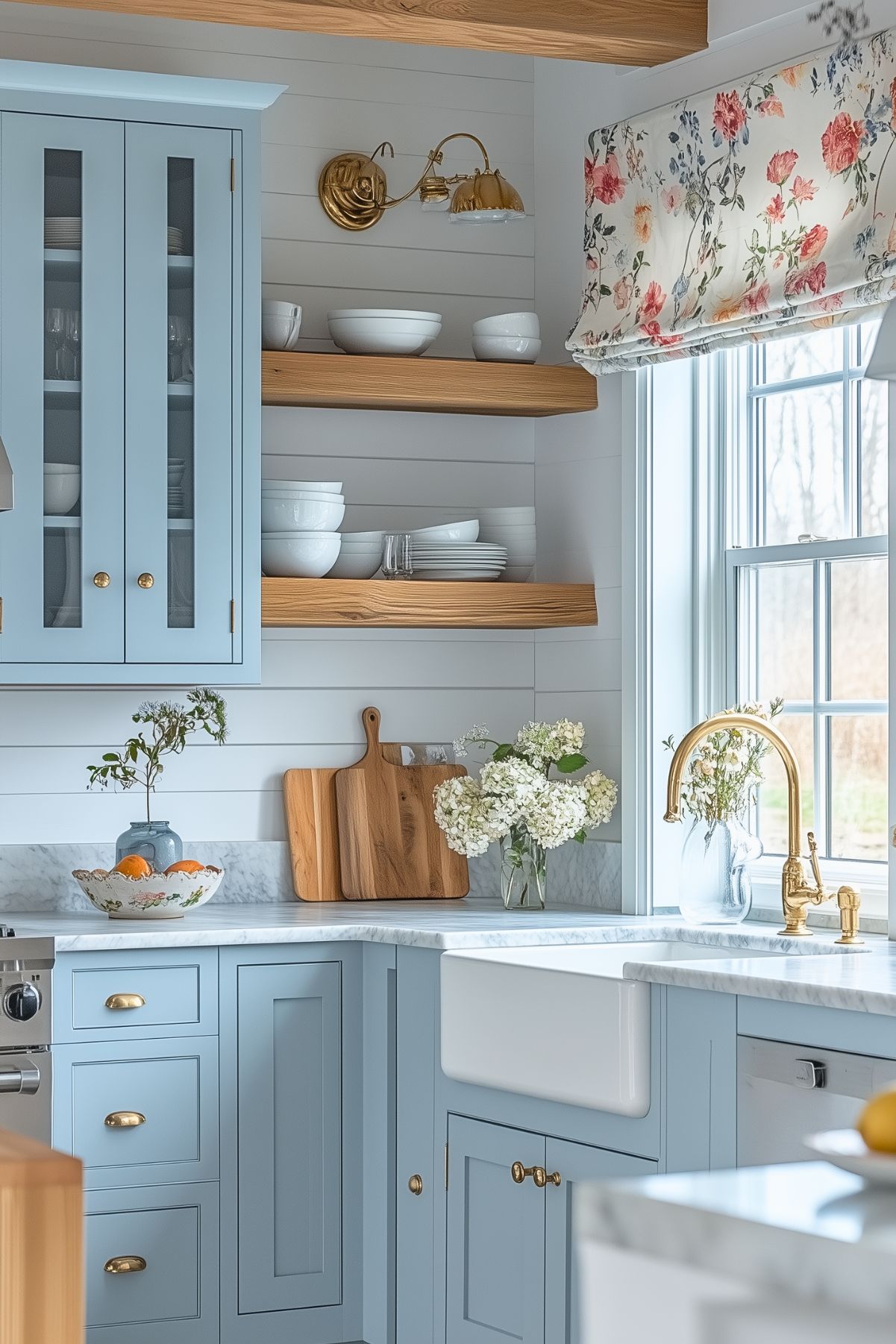 A charming kitchen with soft blue cabinetry, featuring gold hardware and a farmhouse sink. The space has open wooden shelves displaying simple white dishes, adding a natural, rustic feel. The countertops are made of marble, complementing the elegant gold faucet. A floral Roman shade adorns the window, bringing a touch of English country charm. Fresh flowers and wooden cutting boards on the counter enhance the cozy and welcoming atmosphere. The overall design blends classic cottage style with modern functionality, creating a serene and stylish space.