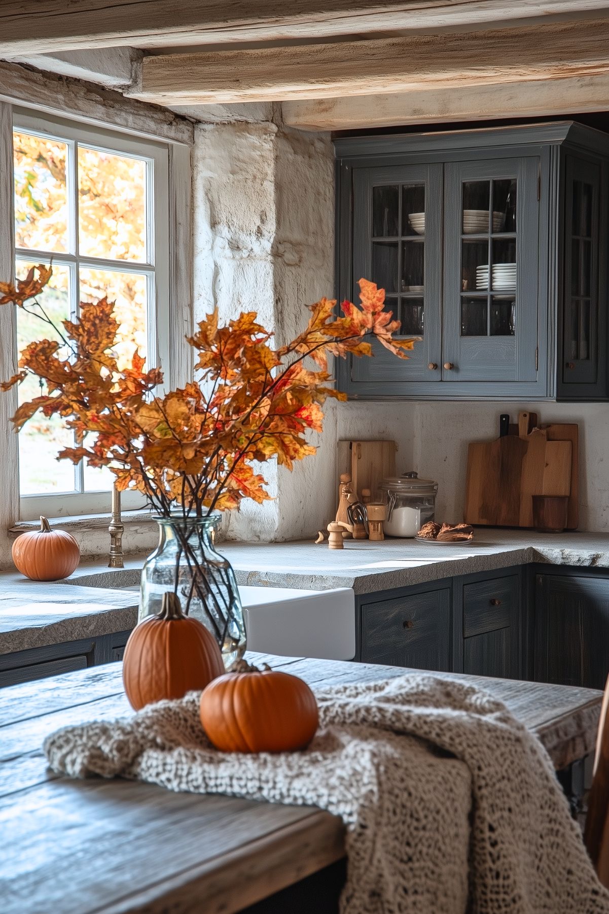 A rustic kitchen decorated for fall with small pumpkins and a vase filled with vibrant orange autumn leaves as the centerpiece. The kitchen features stone countertops and distressed wood cabinetry, enhancing the cozy, country feel of the space. A knitted blanket draped over the table adds warmth to the setting, while cutting boards and kitchen essentials are neatly arranged on the counter. Natural light streams in through the window, illuminating the autumnal decor and creating a welcoming ambiance.