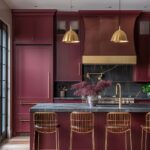 A contemporary kitchen with maroon cabinetry, brass accents, and a dark grey countertop. Two brass dome pendant lights hang over the kitchen island, which is paired with brass wire bar stools. The backsplash is black marble with white veining, and a brass pot filler is mounted above the stove. A large vase filled with dried flowers sits on the island, adding a decorative touch. Light wooden flooring contrasts with the darker tones of the cabinetry and countertops.