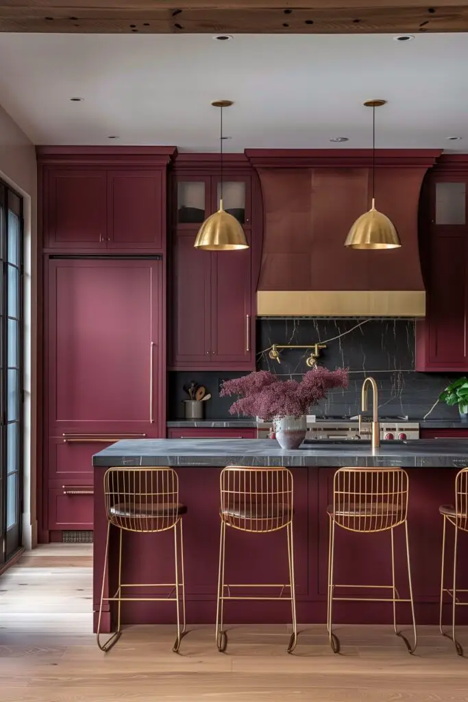 A contemporary kitchen with maroon cabinetry, brass accents, and a dark grey countertop. Two brass dome pendant lights hang over the kitchen island, which is paired with brass wire bar stools. The backsplash is black marble with white veining, and a brass pot filler is mounted above the stove. A large vase filled with dried flowers sits on the island, adding a decorative touch. Light wooden flooring contrasts with the darker tones of the cabinetry and countertops.