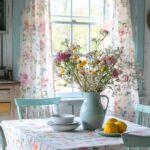 A bright and airy kitchen scene featuring a table covered with a floral tablecloth. The table is set with a light blue pitcher holding a bouquet of colorful wildflowers, a stack of white bowls, and a plate of lemons. The background shows a window with floral curtains that allow natural light to stream in, enhancing the fresh and cheerful cottagecore ambiance of the space.