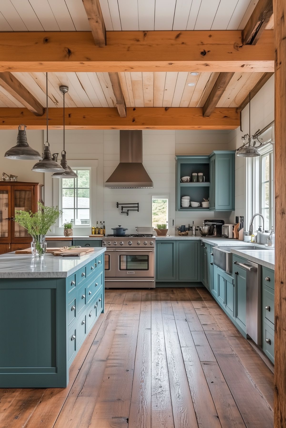 A rustic kitchen with teal-blue cabinetry, stainless steel appliances, and exposed wooden beams on the ceiling. The room features a large island with a marble countertop and farmhouse sinks. The natural wood flooring adds warmth to the space, while industrial-style pendant lights provide an elegant touch.