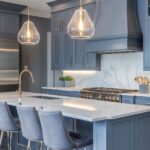 A modern kitchen with blue-gray cabinetry, marble countertops, and elegant glass pendant lights hanging above the island. The kitchen island features a built-in sink and seating with plush blue bar stools. A marble backsplash and gold accents complete the sophisticated look of the space.