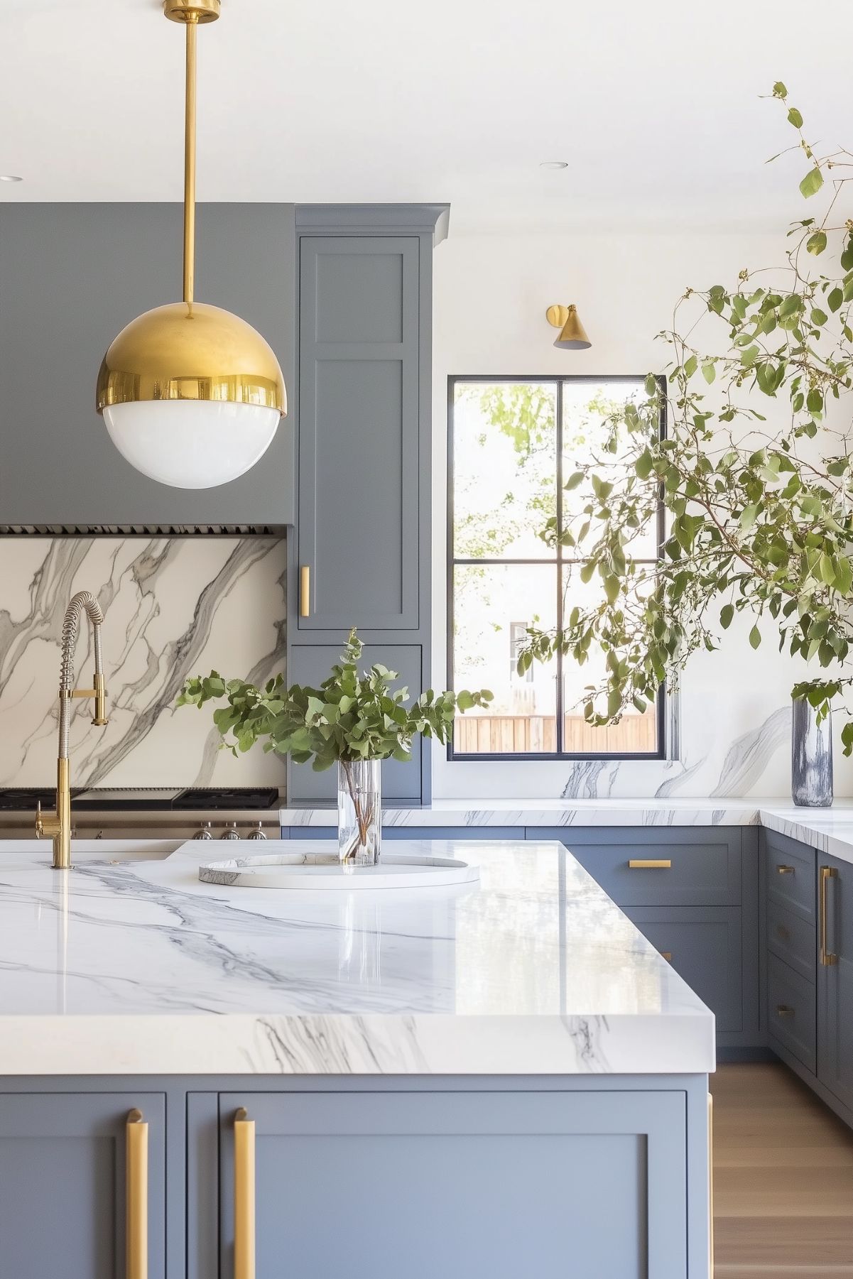 A bright kitchen with blue-gray cabinets, white marble countertops, and gold hardware. A gold pendant light hangs above the island, and a marble backsplash adds an elegant touch. The space is decorated with greenery in a clear vase, giving the kitchen a fresh and modern feel.






