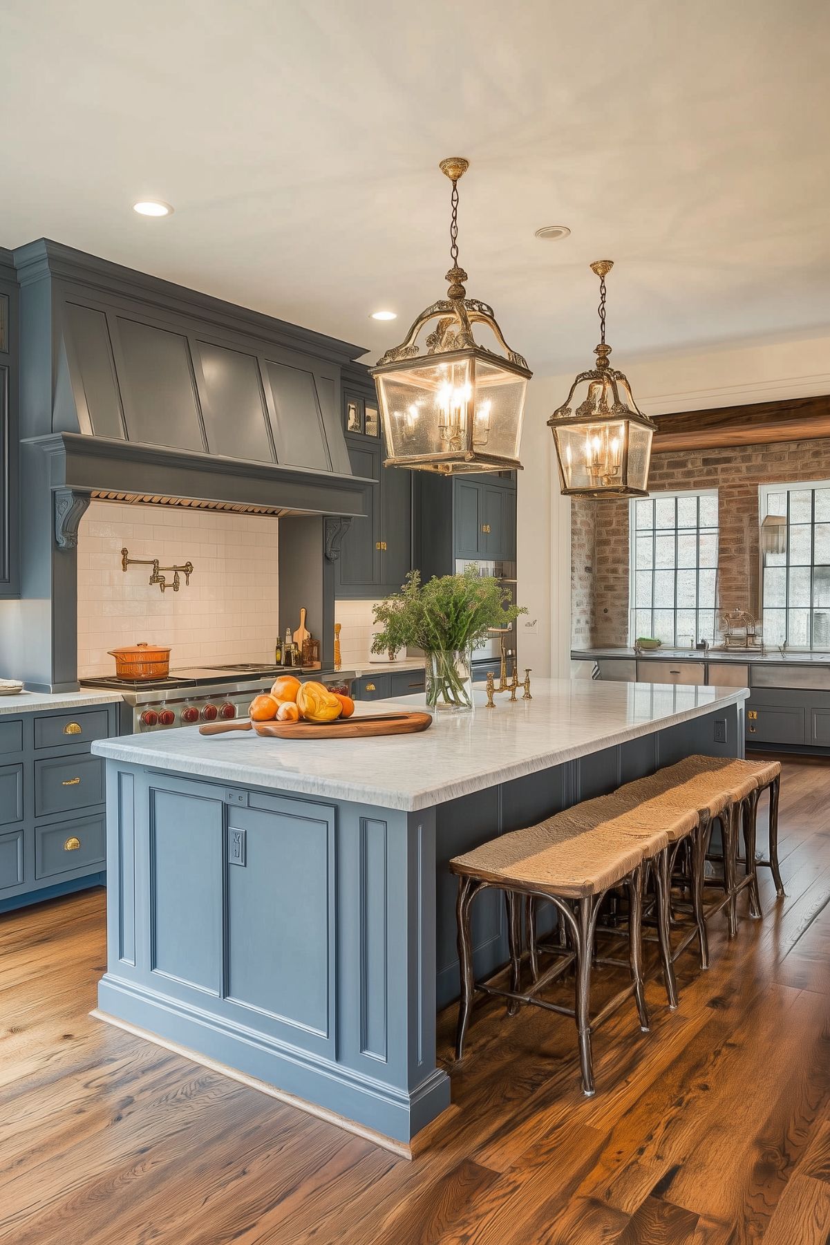 A traditional kitchen with blue-gray cabinetry, a large marble island, and wicker barstools. The space features elegant brass fixtures, two large lantern-style pendant lights, and exposed brick accents. The kitchen island is decorated with fresh fruit and greenery, adding a natural, warm touch to the space.