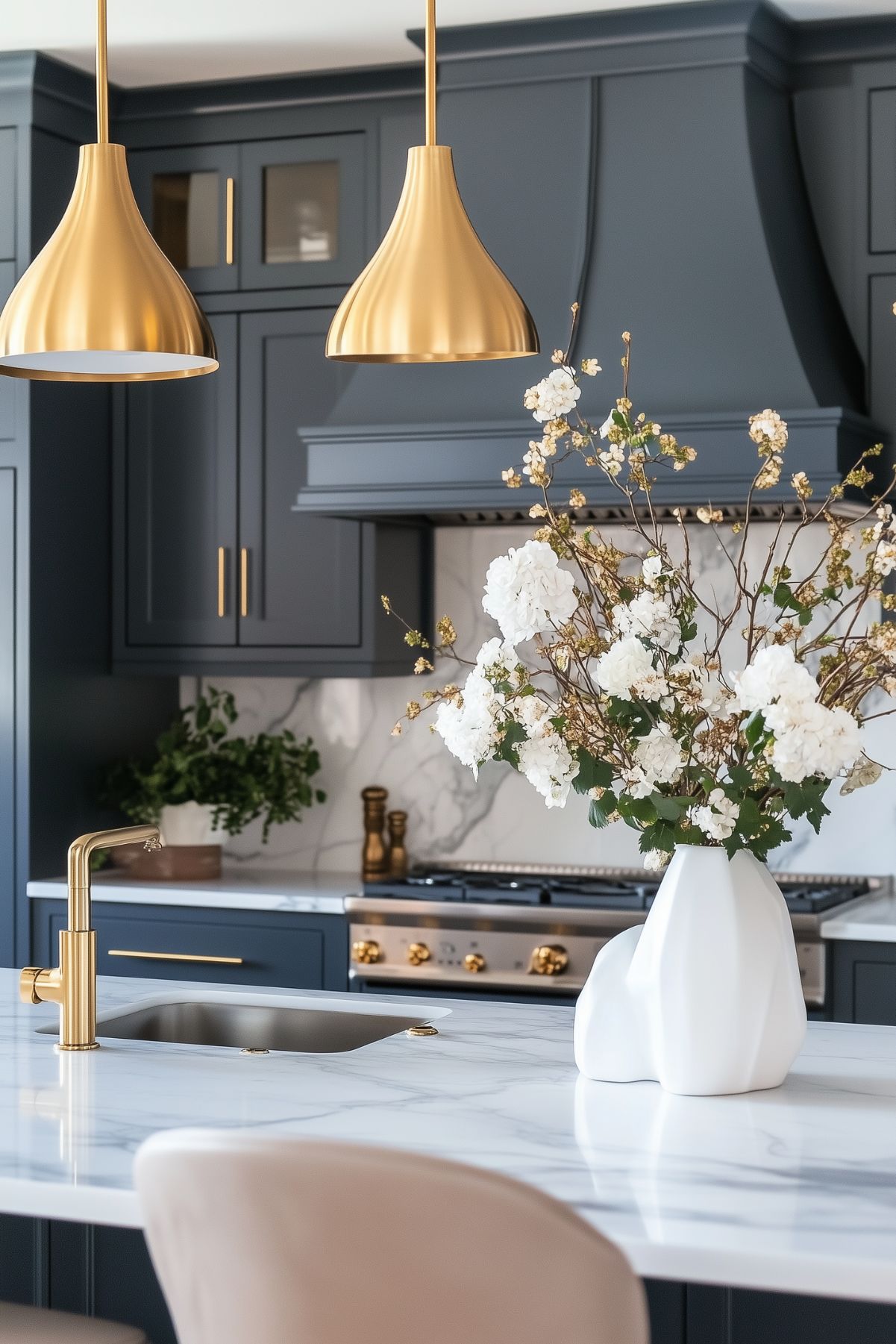 A modern kitchen with blue-gray cabinets, gold pendant lights, and a marble countertop. The island features a gold faucet and a white vase with white flowers as a centerpiece.