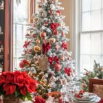 A Christmas tree decorated with red bows, gold and red ornaments, and topped with a gold star is placed in a dining room next to a window. The tree is flocked with artificial snow. A dining table is set in front of the tree, featuring plates, crystal glasses, silverware, and a centerpiece with poinsettias and pinecones, all arranged on a red velvet table runner.