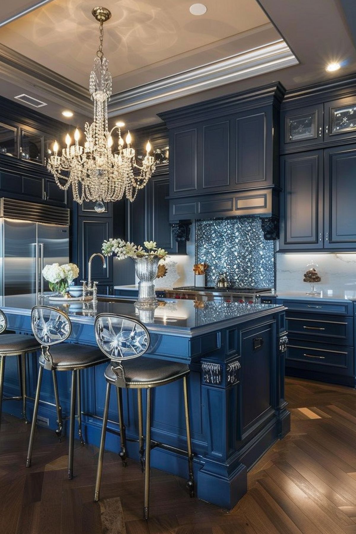 A stunning kitchen with rich navy blue cabinetry and a matching island topped with a glossy countertop. The room is illuminated by a large crystal chandelier, adding elegance. Transparent acrylic barstools with gold frames complement the space, while a mirrored mosaic backsplash enhances the luxurious feel.