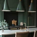 A modern kitchen with dark green cabinetry and a sleek design. Three cone-shaped pendant lights with brass accents hang above a marble kitchen island. The island features a brass faucet and a pair of leather barstools. In the background, a black and gold stove stands out against the dark green cabinetry, and wooden cutting boards add a touch of warmth. A vase with eucalyptus branches adds a subtle decorative touch, completing the sophisticated and stylish look.