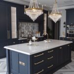 An elegant navy blue kitchen featuring a large island with white countertops and gold hardware. Two crystal chandeliers hang above the island, adding a luxurious touch. The backsplash showcases an intricate tile design, while the marble-patterned floor enhances the sophisticated atmosphere.