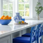 A bright kitchen scene featuring a white marble countertop with a blue vase, a blue bowl of oranges, and blue bar stools. The backdrop shows large windows with patterned blue-and-white curtains, allowing natural light into the space.