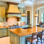 Traditional kitchen with sage green cabinetry, a large gold granite island, and ornate pendant lighting. A custom range hood and intricate wood details add a touch of elegance.