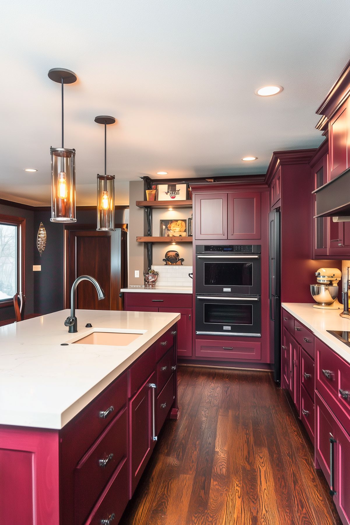 A sophisticated kitchen featuring deep burgundy cabinetry and an island with a white countertop and built-in sink. The rich color of the cabinets contrasts beautifully with the white surfaces, while dark wood floors add warmth and depth. Open wooden shelves on the side wall display decorative items, enhancing the kitchen's rustic charm. Industrial-style pendant lights with glass cylinders and exposed bulbs hang above the island, casting a warm glow. Stainless steel appliances, including double ovens, are seamlessly integrated into the cabinetry, adding a modern touch to this elegant, inviting space.