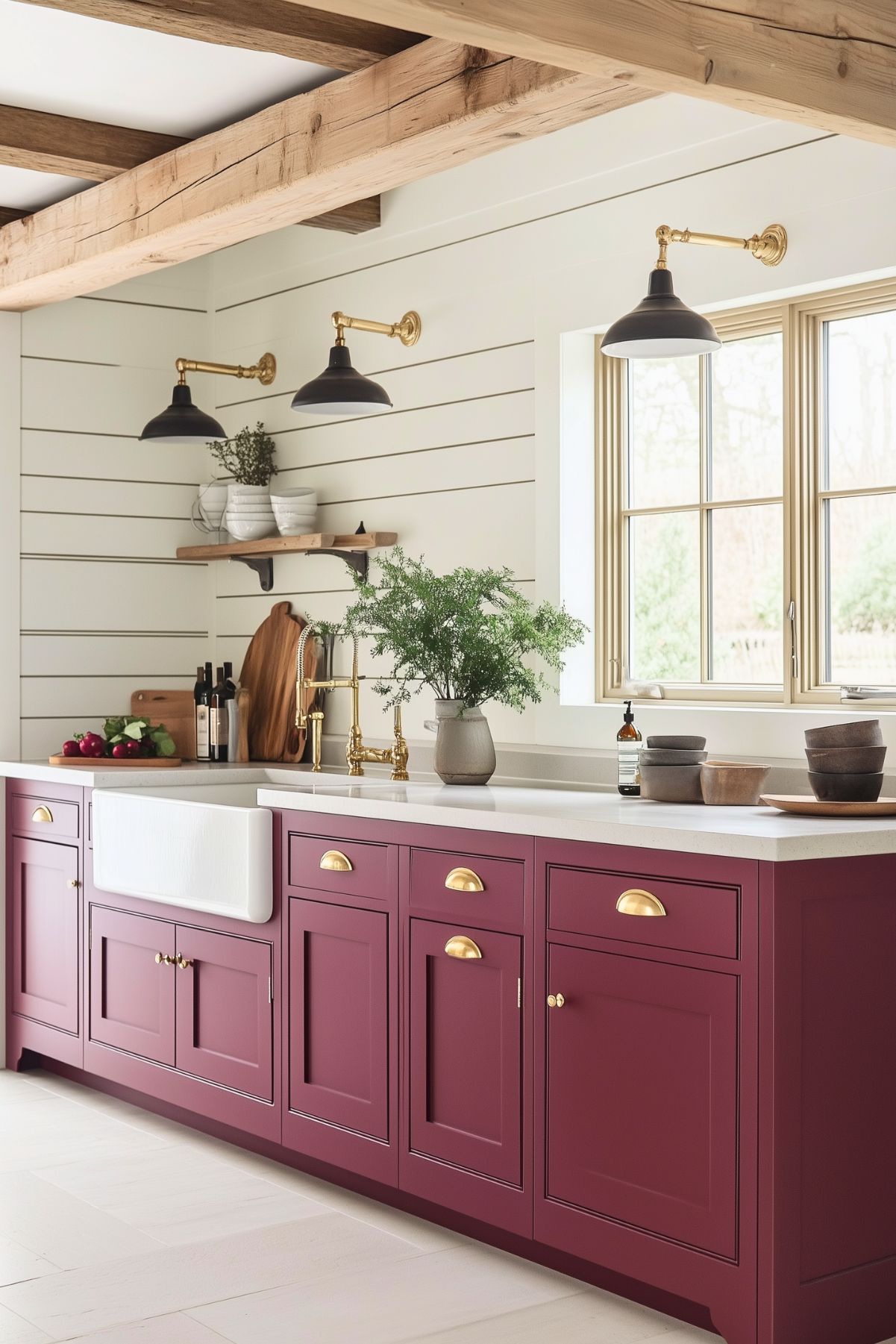 A charming farmhouse kitchen with a modern twist, featuring deep burgundy cabinetry with brass cup pulls, adding warmth and elegance to the space. The white quartz countertop complements the burgundy cabinets, while a large farmhouse apron sink sits in the center, paired with a vintage-inspired brass faucet. Above the sink area, white shiplap walls and open wooden shelves display rustic ceramic dishes and cutting boards, enhancing the farmhouse aesthetic. Black wall-mounted sconces with brass accents provide ambient lighting, and natural wood beams on the ceiling add a touch of rustic warmth. A vase with greenery on the counter brings a fresh element into the space.