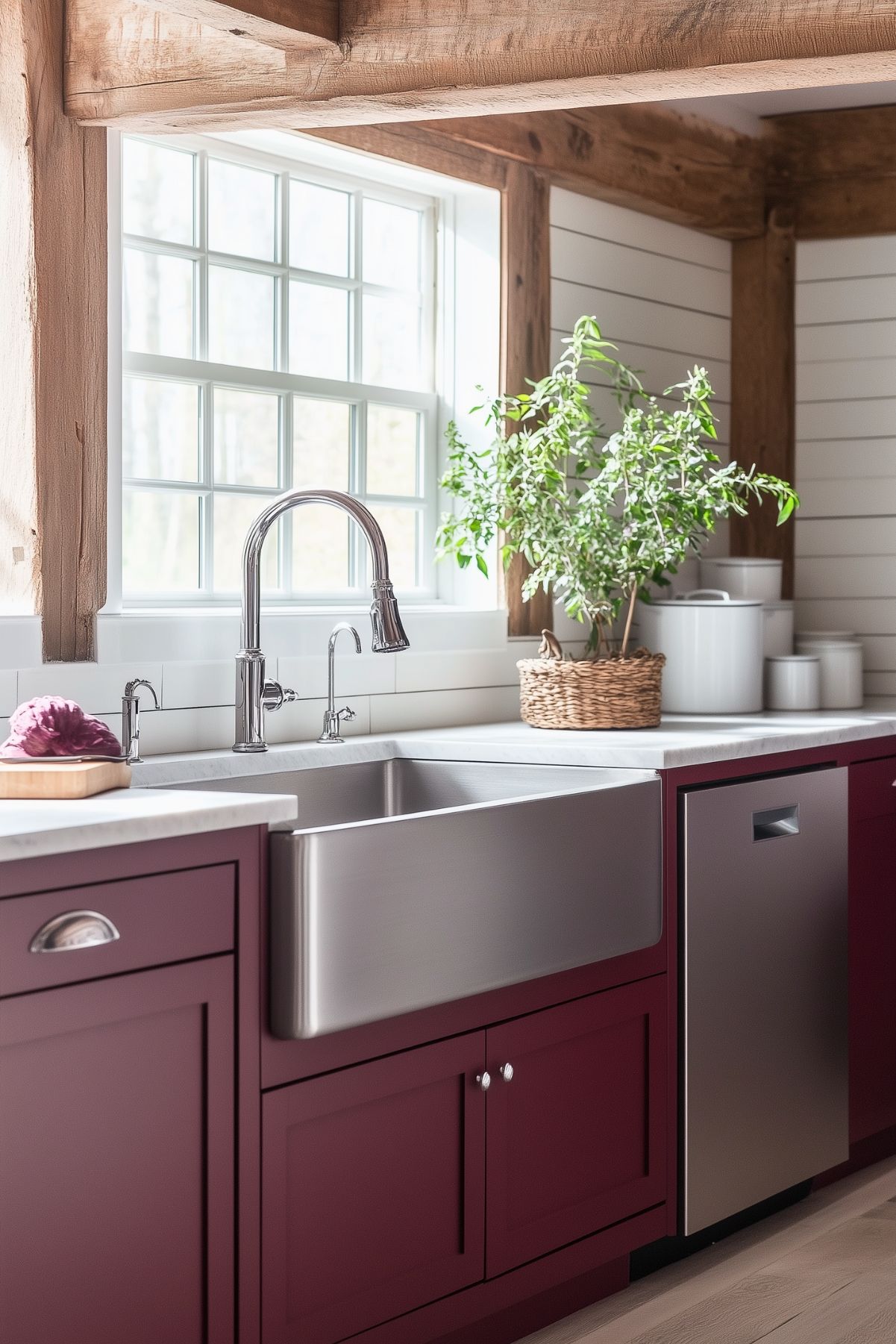 A farmhouse kitchen featuring deep burgundy lower cabinets topped with a sleek white countertop. The focal point is a large stainless steel farmhouse sink with a high-arched gooseneck faucet and additional side sprayer. Above the sink, a window framed by rustic wooden beams brings in natural light, highlighting the shiplap walls. To the side, a woven basket with lush greenery adds a touch of nature, while a collection of white ceramic canisters provides extra storage and complements the clean, rustic aesthetic. The combination of warm wood, rich burgundy, and stainless steel gives this kitchen a cozy yet modern farmhouse feel.