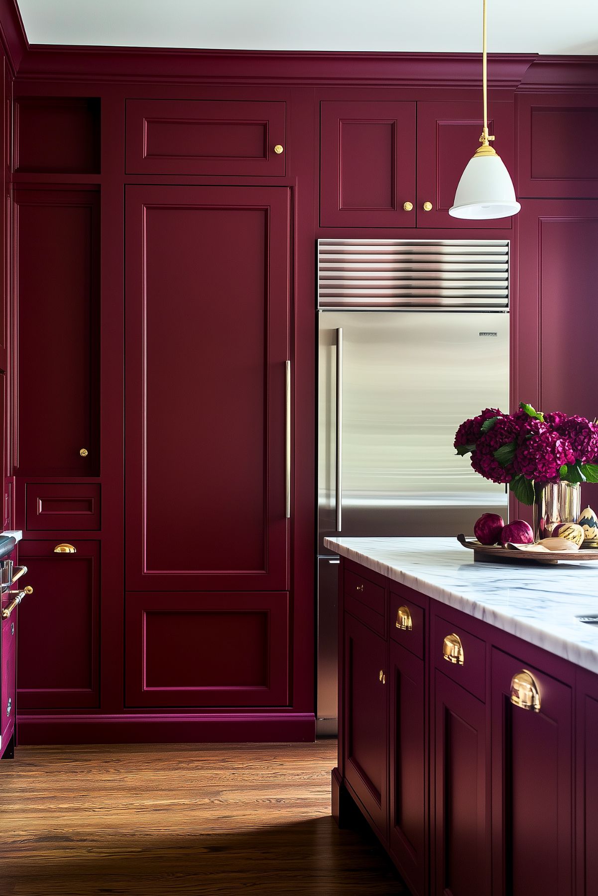 A sophisticated kitchen featuring deep burgundy cabinetry with brass accents, creating a rich and timeless look. The custom cabinets reach the ceiling, enhancing storage space and giving the kitchen a refined aesthetic. A large stainless steel refrigerator is built seamlessly into the cabinetry for a cohesive appearance. The island, topped with a luxurious white marble countertop with subtle gray veining, provides ample workspace. A gold pendant light with a white shade hangs above, adding elegance to the space. A vase of deep magenta flowers and fresh produce on the counter add a pop of natural color.